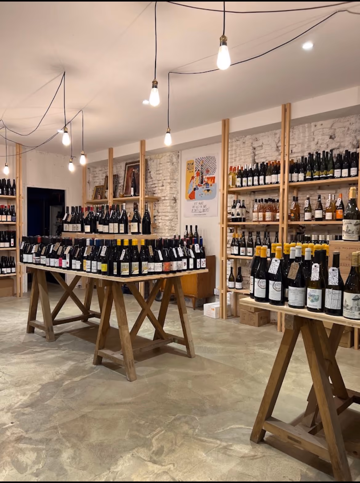 La cave : ampoules nues, tréteaux en bois couverts de bouteilles, murs de brique blanchie