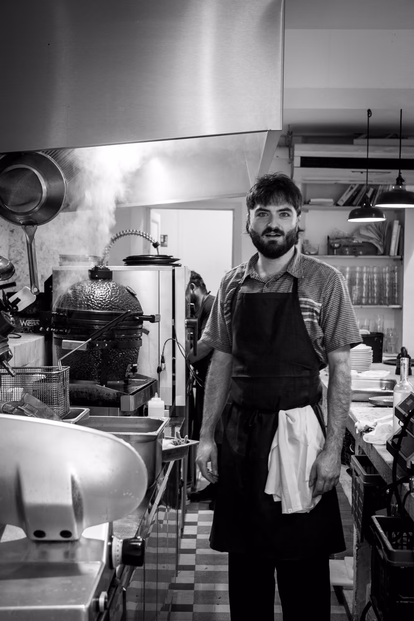 Tanguy, le chef de Midi Minuit, debout dans la cuisine, torchon au tablier, en noir et blanc