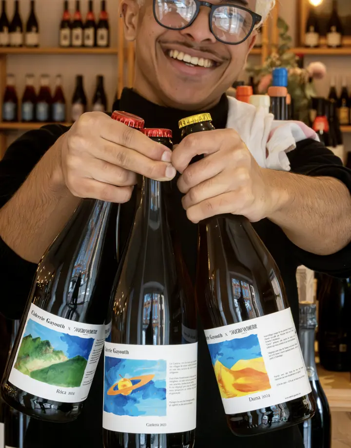 Un sourire et trois bouteilles de cidre présentées devant les étagères de la cave